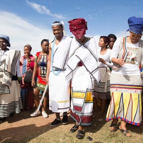 A group of people, as if stepping right out of a Lesedi Cultural Village Tour, don traditional African attire during a vibrant cultural celebration under the blue sky. Adorned with colorful patterns and headpieces, they walk together on the lush grass, showcasing their rich heritage.