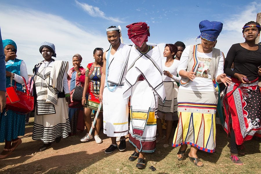 A group of people, as if stepping right out of a Lesedi Cultural Village Tour, don traditional African attire during a vibrant cultural celebration under the blue sky. Adorned with colorful patterns and headpieces, they walk together on the lush grass, showcasing their rich heritage.