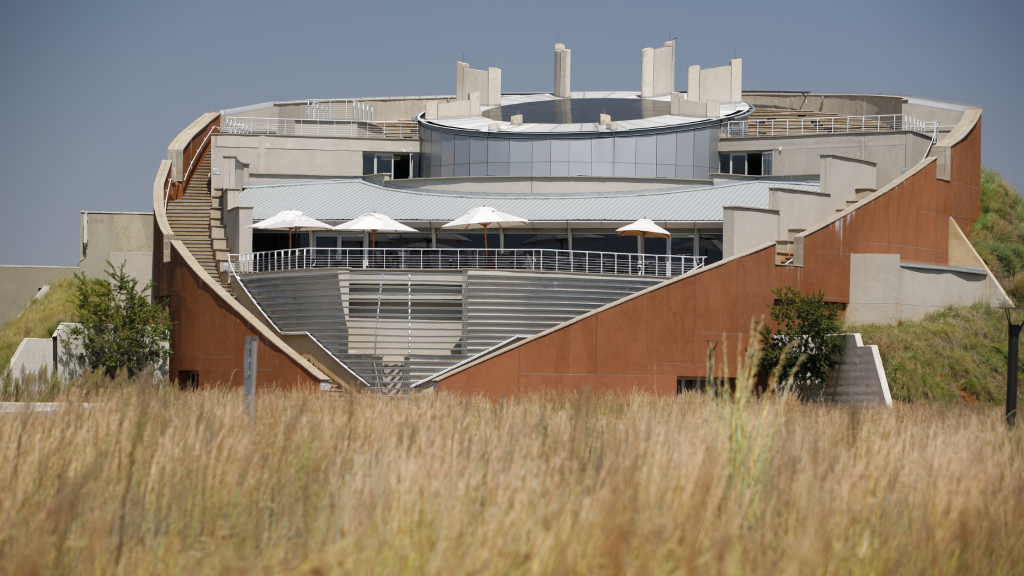 A modern, circular building with a stepped design and large windows stands amid tall grass, resembling the architectural marvels highlighted on a Cradle of Human Kind Day Tour. The structure features a rooftop terrace with umbrellas against the backdrop of a clear blue sky.