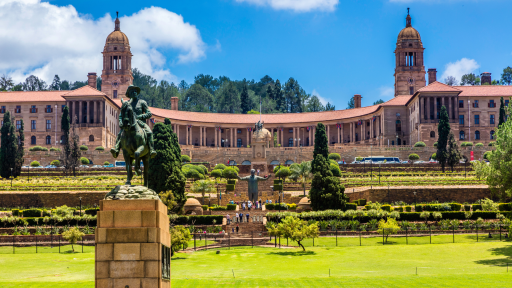 Experience the panoramic view on a Pretoria City tour, showcasing a large historic building with domed towers, surrounded by gardens and a statue of a rider on horseback in the foreground. The clear blue sky and scattered trees enhance this picturesque landscape.