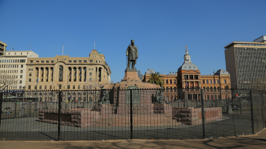 A statue of a man stands atop a large pedestal, surrounded by smaller statues in a fenced area. This iconic landmark is a highlight of the Pretoria city tour, with historic buildings featuring classic architectural styles under the clear blue sky as its stunning backdrop.