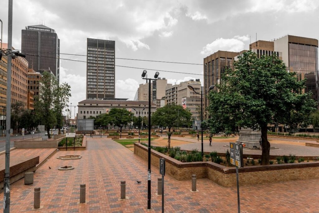 A city plaza featuring brick pathways, green trees, and surrounding tall buildings under a cloudy sky evokes the essence of an urban Soweto tour. The area is equipped with streetlights and signage, blending the city's vibrant history with a touch of nature.