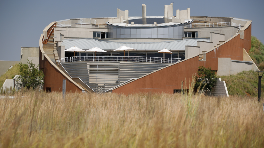 A modern, circular building with a layered design, featuring large terraces and multiple white umbrellas, evokes the spirit of a Lesedi Cultural Village tour. Surrounded by tall, dry grass under a clear blue sky, it harmonizes with the essence of the Cradle of Humankind.