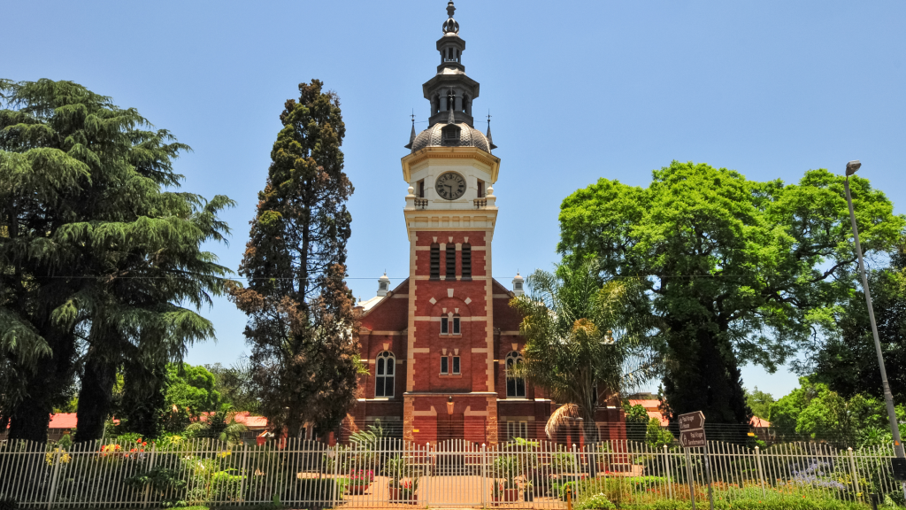 A historic red brick church with a clock tower and ornate detailing stands proudly against Pretoria's clear blue sky. Lush green trees envelop the building, while a metal fence runs along the foreground, making it a must-see on any Pretoria city tour.