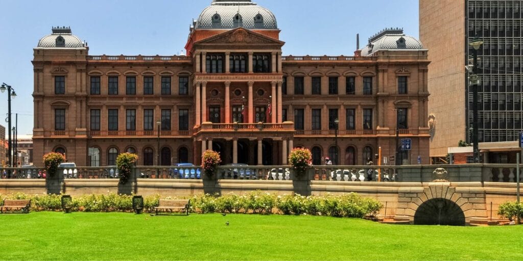 A grand, historic building with intricate architectural details and a central dome stands against a clear blue sky, reminiscent of landmarks seen on a Soweto and Apartheid Museum tour. In the foreground, there's a well-maintained lawn with flowerbeds and benches, bordered by a stone wall.