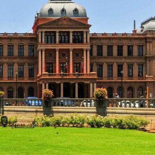 A grand, historic building with intricate architectural details and a central dome stands against a clear blue sky, reminiscent of landmarks seen on a Soweto and Apartheid Museum tour. In the foreground, there's a well-maintained lawn with flowerbeds and benches, bordered by a stone wall.