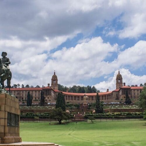 A statue of a man on horseback graces the foreground of a large green lawn. In the background, reminiscent of a Soweto landscape, stands a grand building with twin towers and a red roof beneath a cloudy sky. Trees line the area, evoking reflections similar to those from an Apartheid museum tour.