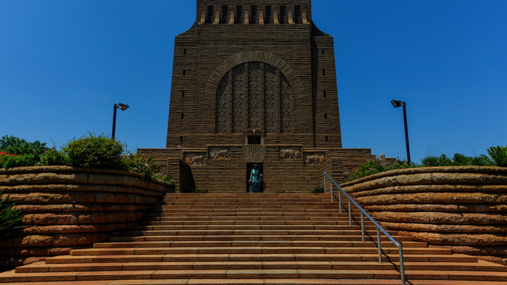 The image captures a highlight of the Pretoria city tour: a large brown stone monument with intricate carvings, surrounded by lush greenery. A wide staircase leads to the entrance, centered by a prominent statue, all set against a bright, clear blue sky.