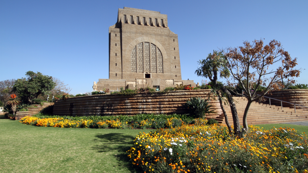 A large stone monument with intricate carvings stands under a clear blue sky, a highlight of the Pretoria city tour. It's surrounded by a circular garden with bright yellow and white flowers. A few trees are scattered around the well-maintained lawn, creating a serene atmosphere.