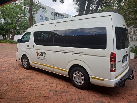 A white passenger van, emblazoned with a logo, is parked on a brick driveway near trees and a building. It's poised for adventure, ready to transfer you to the Pilanesberg National Park.