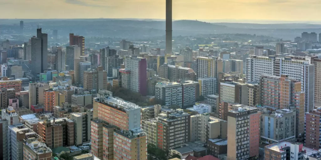 Aerial view of a sprawling cityscape with numerous high-rise buildings under a hazy sky. In the background, hills can be seen with a tall tower prominently rising above the skyline.