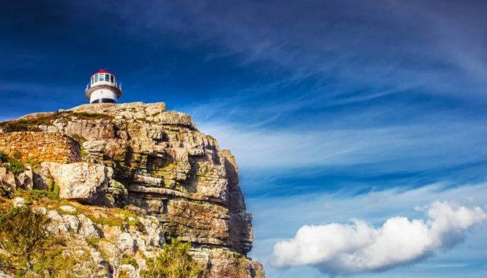 Cape of Good Hope A lighthouse perched atop a rugged cliff under a vivid blue sky with wisps of cloud, much like the scenic views on a Cape Peninsula tour. The landscape is dotted with sparse greenery, conveying solitude and natural beauty.