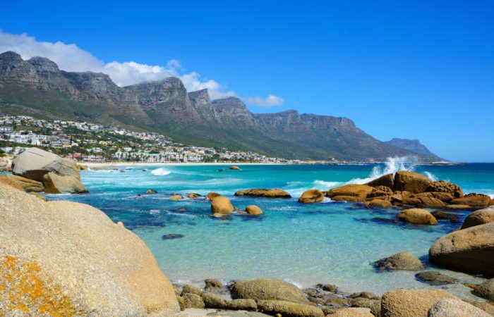 camps bay Experience the Best of the Cape Tour along a rocky shoreline with turquoise waters where waves crash against boulders. Rugged mountains rise in the distance under a clear blue sky, while sunlit, white buildings pepper the vibrant hillsides.