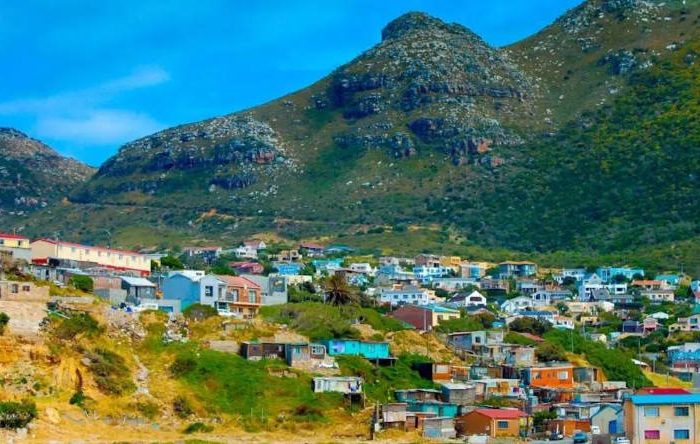 HOUT BAY Colorful houses are scattered across a hillside, with lush green mountains in the background under a clear blue sky. Embark on a private Cape Peninsula tour to experience this vibrant community that seamlessly blends with the natural landscape, creating a picturesque scene.