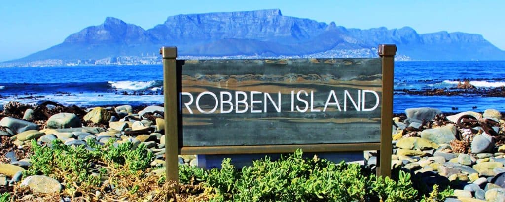 A sign reading Robben Island stands amid rocky terrain and green plants, offering a glimpse into the iconic Robben Island and Table Mountain tour. In the background, a body of water and majestic mountain range stretch beneath a clear blue sky.