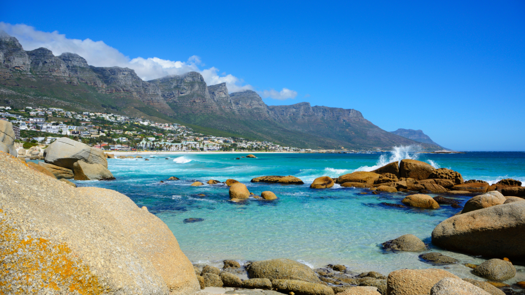 Experience the Best of the Cape Tour along a rocky shoreline with turquoise waters where waves crash against boulders. Rugged mountains rise in the distance under a clear blue sky, while sunlit, white buildings pepper the vibrant hillsides.