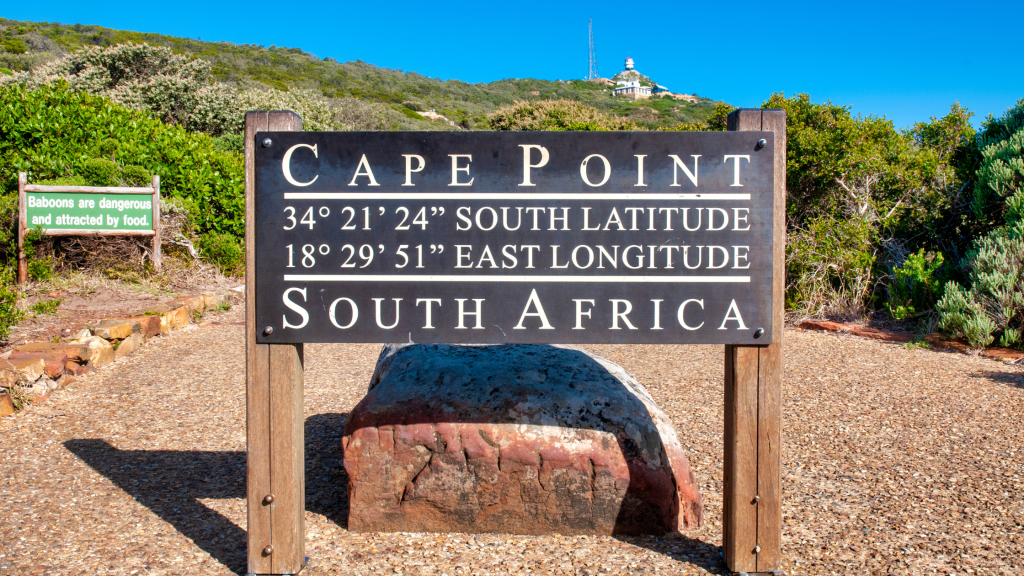 A sign at Cape Point, a highlight of the Cape Peninsula tour in South Africa, displays coordinates: 34°2124 South Latitude and 18°2951 East Longitude. The surrounding landscape boasts lush greenery, a winding path, and a hill crowned with a quaint structure.