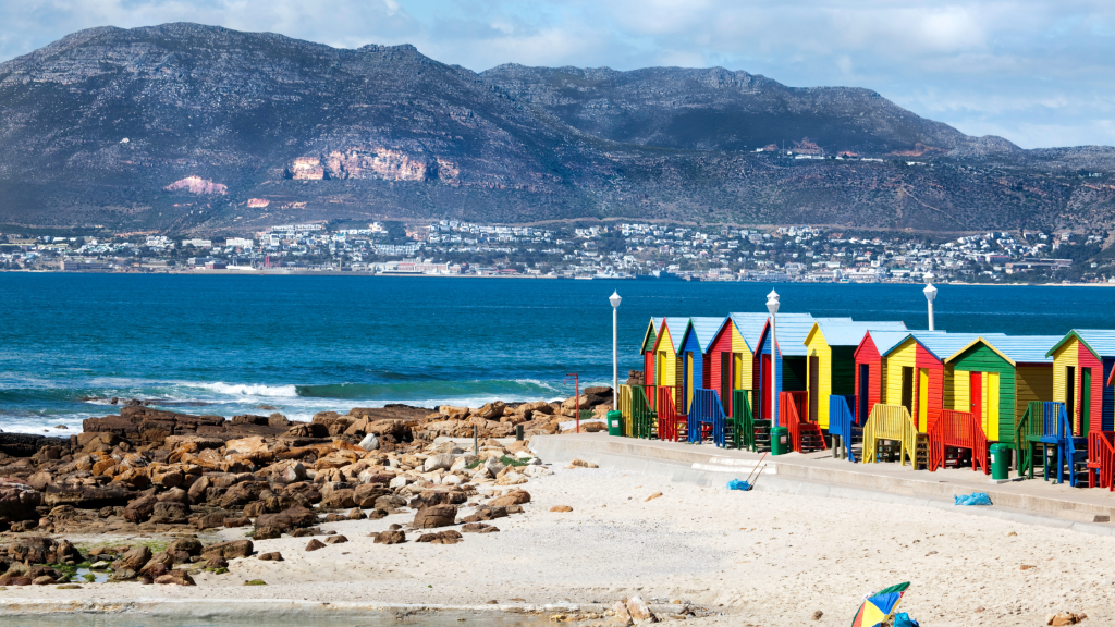 Colorful beach huts line a sandy coastline with rocky patches, set against a backdrop of blue ocean waves and a mountainous landscape. As part of the Cape Peninsula tour, the town is visible across the water under a cloudy sky.