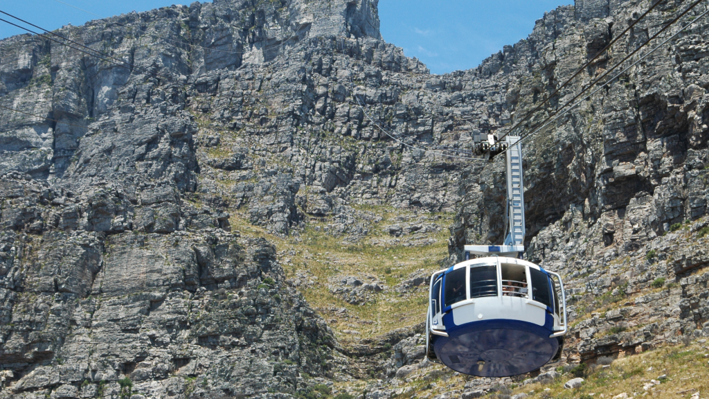 A blue and white cable car, part of the scenic Table Mountain tour, ascends a steep, rocky mountainside under a clear blue sky. The rugged terrain is covered in sparse vegetation.