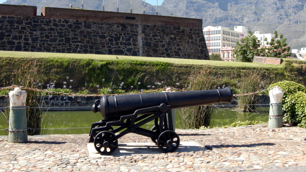 A historic black cannon on a stone platform with wheels is displayed in an outdoor area, surrounded by greenery and a stone fortress wall. The setting captures the essence of a Robben Island and Table Mountain tour, with a backdrop featuring a modern building and mountain under clear blue skies.