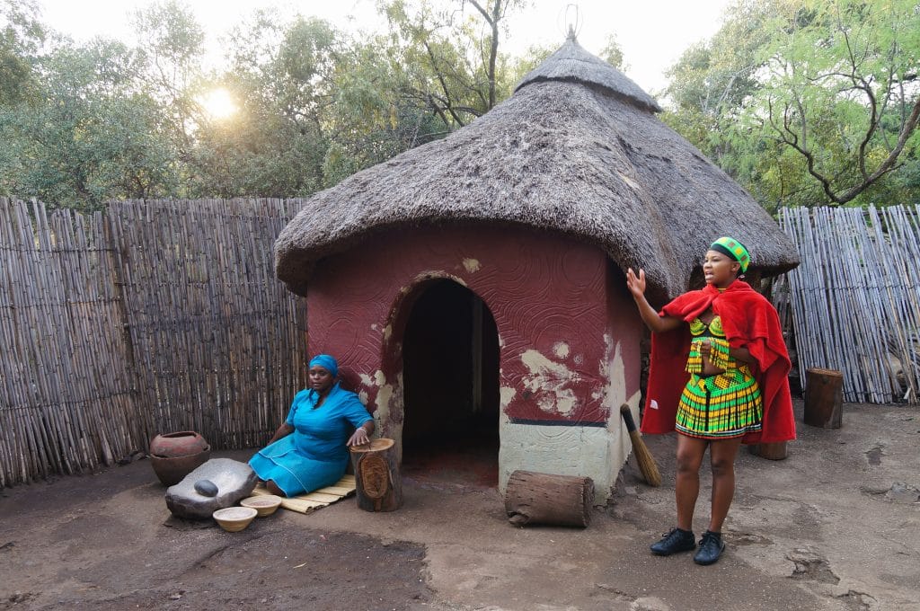 Two people in traditional attire are in front of a round thatched-roof hut, capturing the essence of South Africa. One sits on a log with objects nearby, while the other leans against the hut. A bamboo fence and lush greenery complete the scene, inviting you into this travel guide to South Africa.