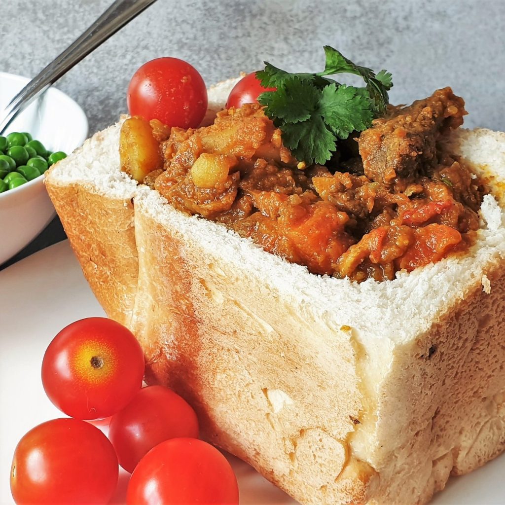 Close-up of a hollowed-out loaf filled with hearty beef stew, reminiscent of flavors highlighted in any travel guide to South Africa. Cherry tomatoes and green peas accompany the dish, garnished with fresh cilantro on a white plate.