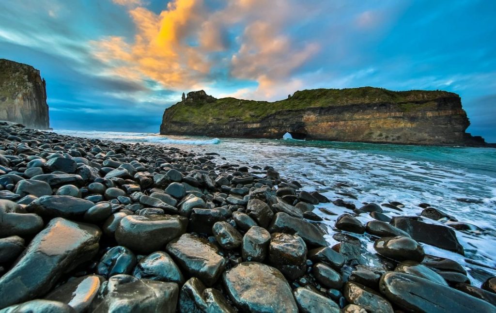 Rocky shoreline with smooth stones leads to a large, grassy rock formation in the ocean. A vibrant sunset with orange and blue hues lights up the sky, reflecting on the water. Waves gently crash against the rocks, creating a serene coastal scene, just like a travel guide to South Africa.