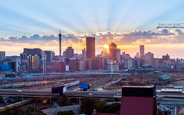 Sunrise over a bustling city skyline with tall buildings, roads, and railway lines paints a perfect scene for your travel guide to South Africa. Sun rays shine through scattered clouds, casting a warm glow on the urban landscape. A construction crane stands on the right, signaling growth and development.