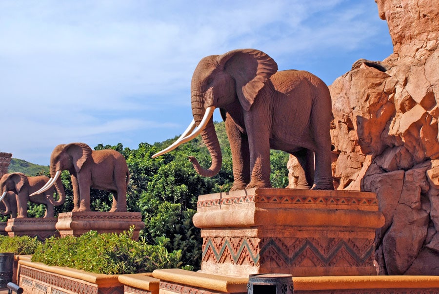 A row of large, detailed elephant statues with long tusks stands on ornate pedestals against a backdrop of rocky terrain and greenery under a clear blue sky, capturing the majestic essence you'd find in any travel guide to South Africa.