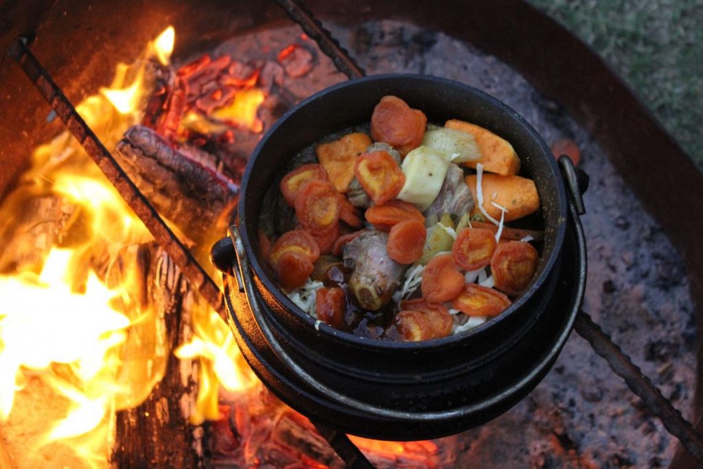 Over an open flame on a grill, a black pot simmers with a hearty South African stew of meat, vegetables, and sliced apricots. This delicious dish might just tempt you to explore more culinary delights from your travel guide to South Africa, where flavors dance like the bright orange flames beneath the pot.