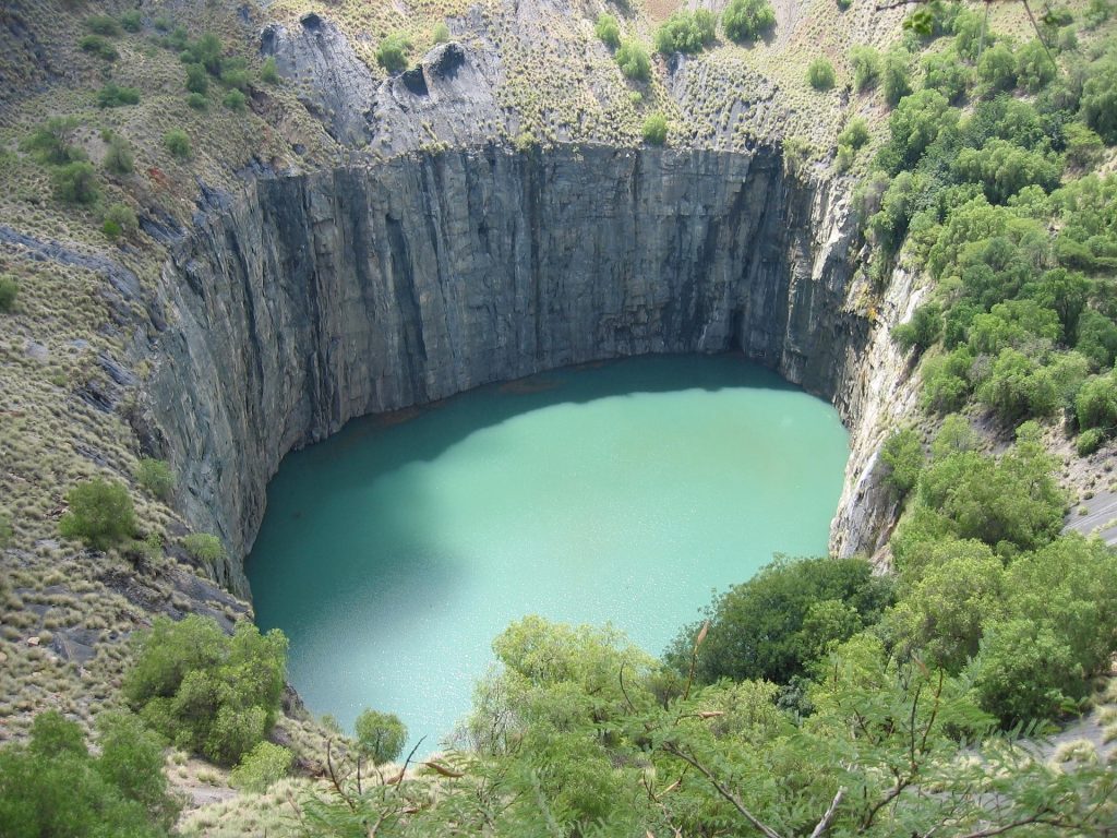 Aerial view of a large, circular pit filled with turquoise water, surrounded by steep, rocky walls in South Africa. The area is lush with green vegetation, creating a stark contrast with the rocky landscape—a must-see for any travel guide to this stunning region.