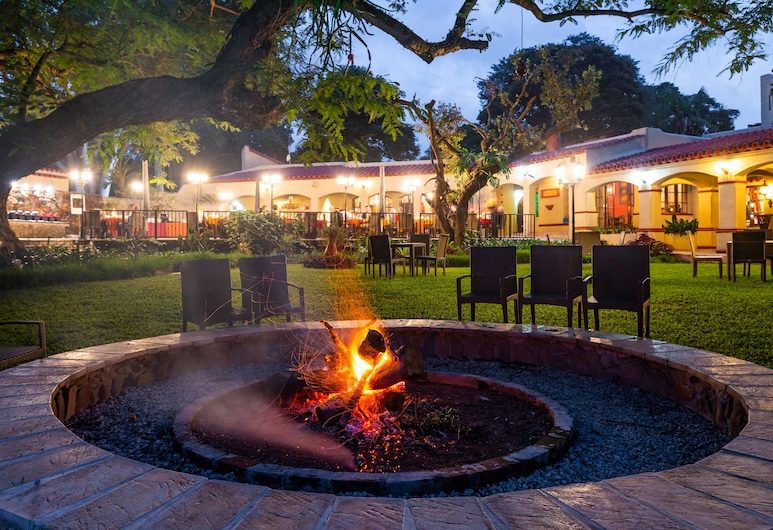 A scenic outdoor area during twilight features a lit fire pit surrounded by chairs. In the background, there's a well-lit building, likely a restaurant or venue, reminiscent of Kruger National Park safari accommodations, with trees and a grassy area adding to the ambiance.