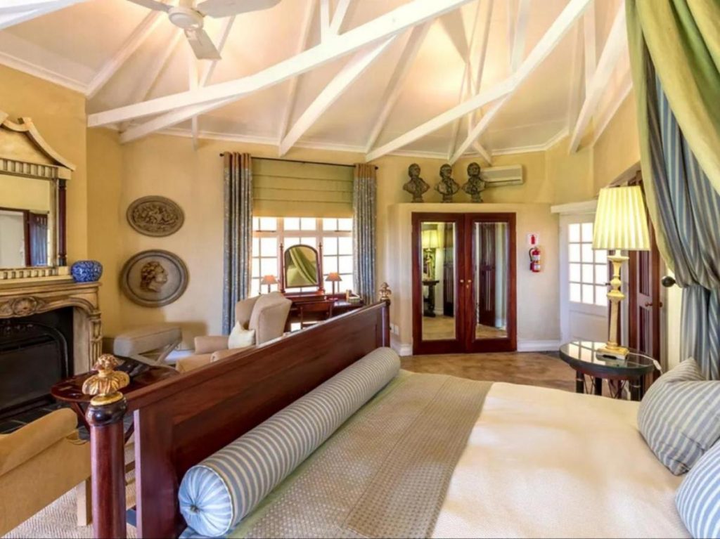 A cozy bedroom with a vaulted ceiling, akin to Kruger National Park Safari Accommodation, features a large bed with striped pillows, a fireplace, mirrors, and chairs. Warm lighting and expansive windows create an inviting ambiance reminiscent of an adventurous yet restful escape.