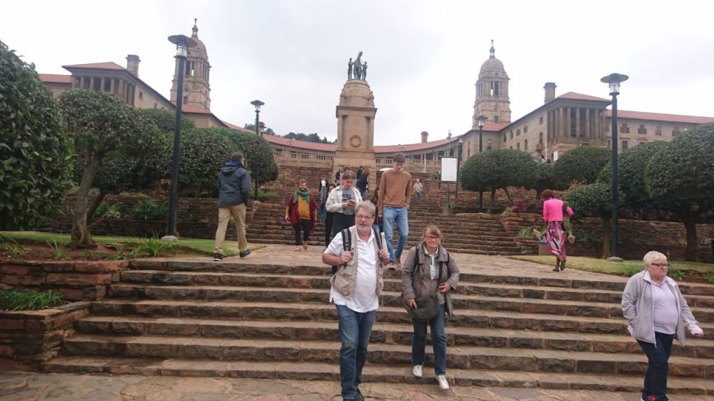 People strolling down stone steps in front of a large historic building in Johannesburg, adorned with domed towers and a central statue. The sky is overcast, and trees and bushes line the path.