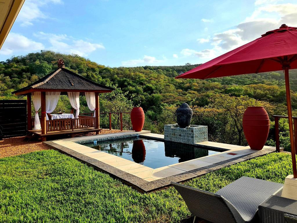 A serene outdoor setting with a thatched gazebo draped in white curtains, overlooking an infinity pool. The accommodation features large red urns and lush greenery, embodying the tranquility of a Kruger National Park safari. A red umbrella shades sun loungers to the right against scenic hills.