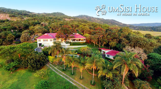 Aerial view of a large house with red roofs, nestled in lush greenery and palm trees near Kruger National Park Safari Accommodation, set against a backdrop of hills and woods. The text "umSisi House At Home in Africa" graces the top right corner, capturing the essence of an African retreat.