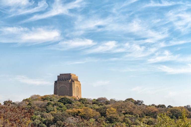 Situated on a grassy hill surrounded by trees, a large stone monument stands as a must-see landmark for anyone embarking on South Africa Overland Tours. The bright sky, dotted with scattered clouds, adds to the tranquil beauty of this serene location.