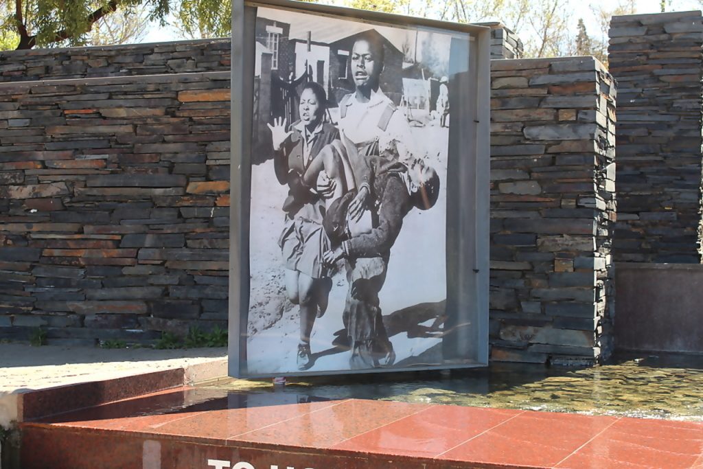 A historical black and white photograph displayed outdoors shows a young man carrying an injured child, with a distressed woman alongside. The background features a stone wall and a partially visible inscription on a red platform.