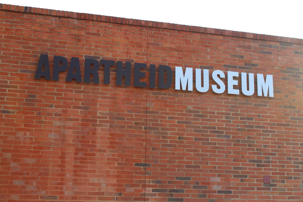 Sign on a red brick wall reads APARTHEID MUSEUM with the words APARTHEID in black and MUSEUM in white.