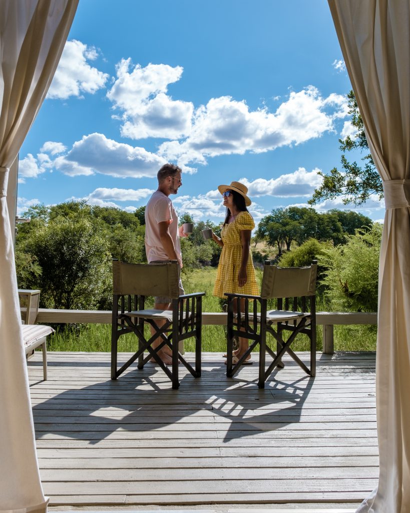 Two people stand on a wooden deck surrounded by nature. They are under a canopy, with two chairs nearby. One person wears a yellow dress and hat, and the other a light shirt and shorts. The sky is blue with scattered clouds.