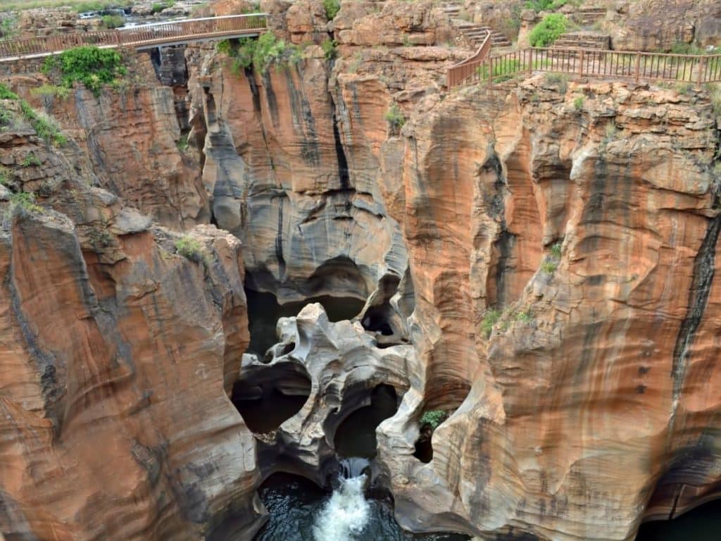 Dramatic rock formations at Bourkes Luck Potholes, featuring deep, cylindrical potholes in reddish-brown rock, with a small stream flowing through. A wooden walkway and green vegetation are visible on the surrounding rocky terrain.