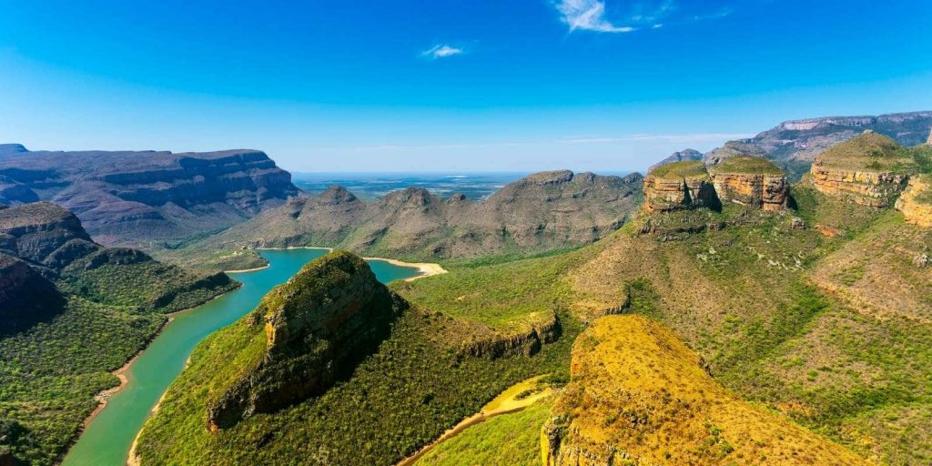 A breathtaking aerial view of a winding river cutting through a vast mountainous landscape with lush greenery under a clear blue sky, creating a stunning natural panorama.