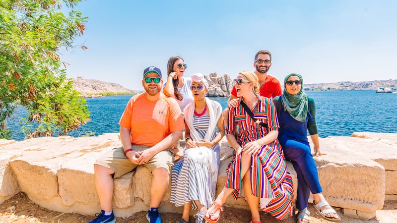 A group of six people sitting on a stone ledge with a scenic backdrop of a large body of water and distant hills. They are casually dressed, smiling, and appear to be enjoying a sunny day. Trees partially frame the left side of the image.