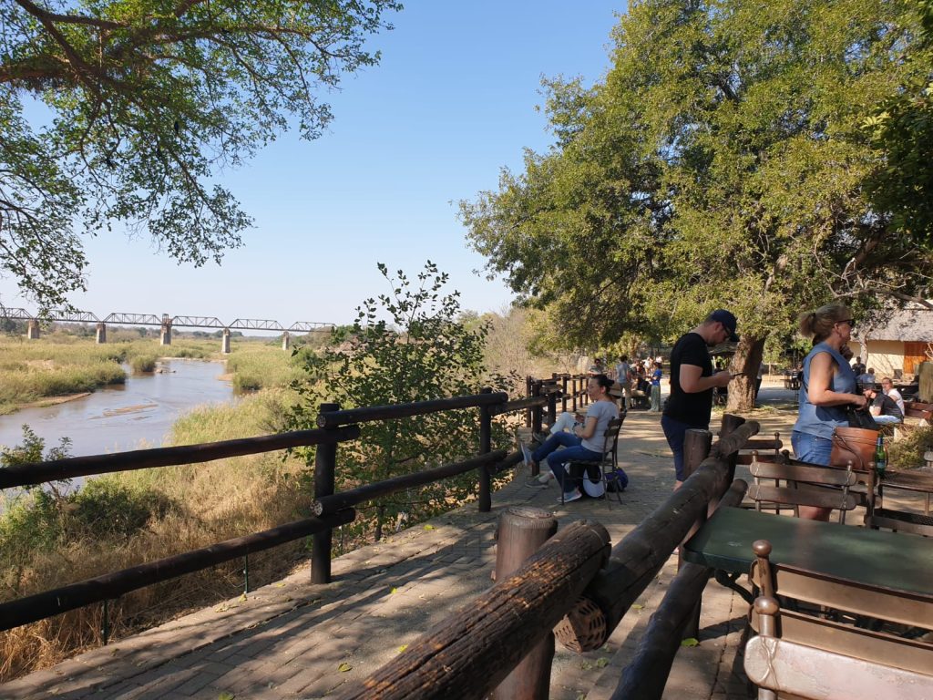 People relax on a patio overlooking a river with a distant metal bridge. The area is shaded by large trees, and the atmosphere is peaceful with clear blue skies. Tables and chairs are arranged along a wooden railing.