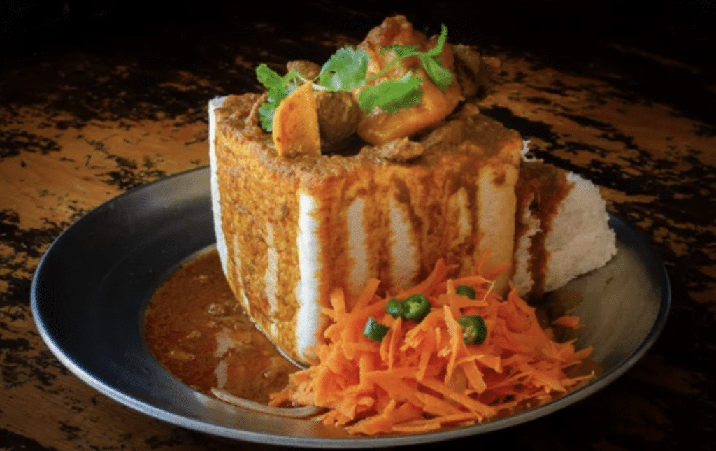 A dish of bunny chow is served on a metallic plate. The hollowed-out loaf of bread is filled with spicy curry and garnished with cilantro. Beside it, there are grated carrots and sliced green chilies. The plate is on a rustic wooden surface.