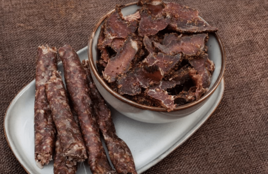 A plate featuring a bowl of sliced dried meat next to several dried sausage sticks, set on a textured brown fabric background.