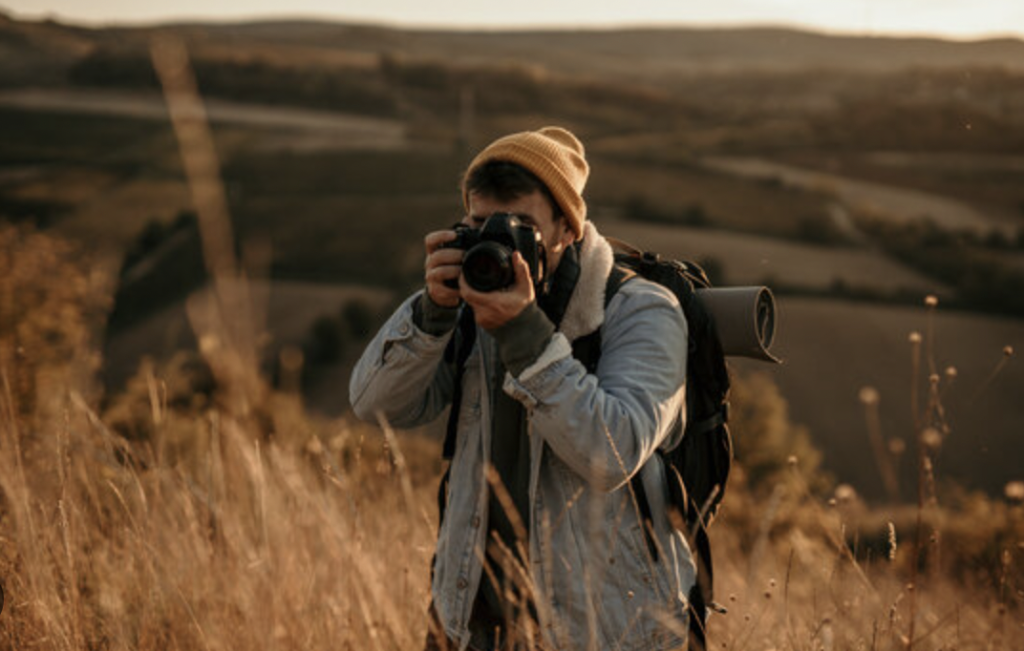 A person wearing a beanie and denim jacket holds a camera up, taking a photo in a golden field. A backpack with a rolled-up mat is slung over one shoulder. The sun is setting over a hilly landscape in the background.