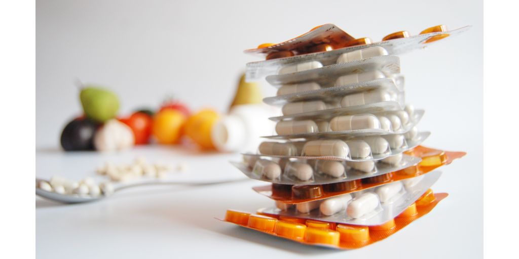 A stack of blister packs containing white pills is in focus, with blurred assorted fruits and a spoon filled with capsules in the background, set against a light gray surface.