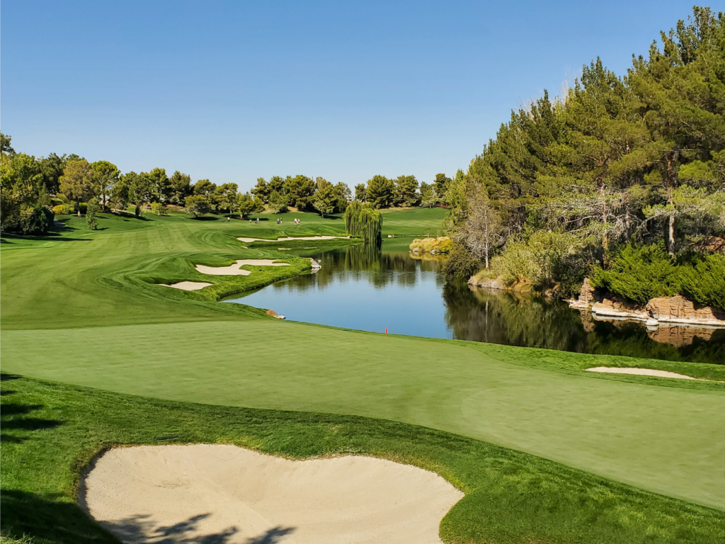 A scenic golf course with a smooth green, several sand traps, and a tranquil water hazard. Trees line the fairway under a clear blue sky, and the landscape gently undulates across the terrain.