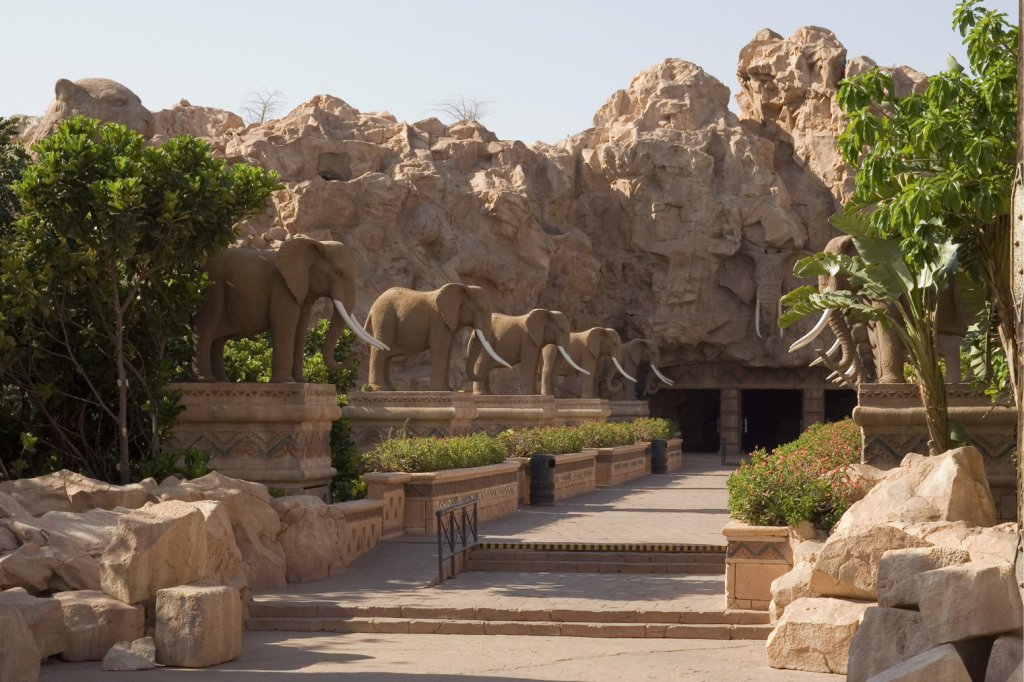 A pathway flanked by elephant statues leads to a rocky cave entrance. Lush green bushes line the walkway, and the background features rugged, sunlit rock formations against a clear sky.